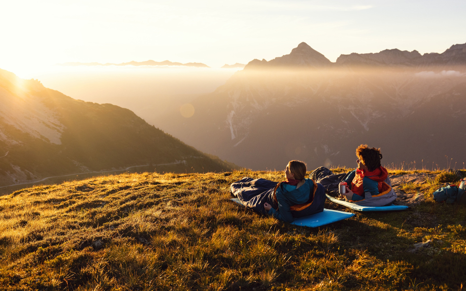 Zwei Wanderer in Schlafsäcken genießen den Sonnenaufgang mit Bergblick auf einer Rucksacktour.