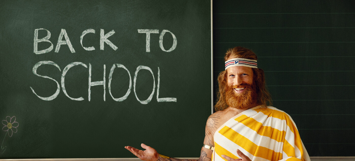 Mann mit Bart in gelb&weiß gestreiftem Handtuch steht vor einer Schultafel auf der BACK TO SCHOOL geschrieben steht.