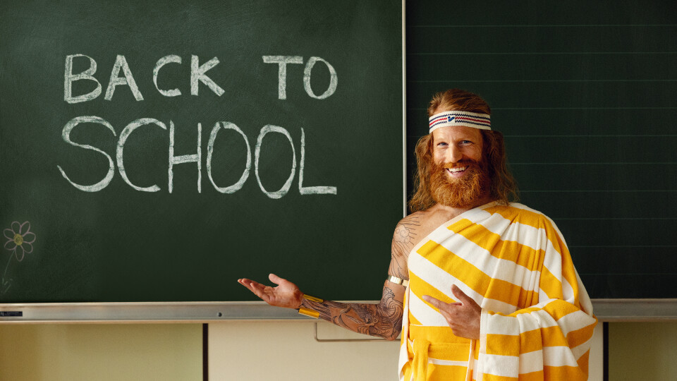 Ein Mann, der den Sommer darstellen soll, steht vor einer Schultafel auf der BACK TO SCHOOL geschrieben ist. Der Mann hat einen Bart, lange Haare und trägt ein weiß, gelbes Handtuch wie eine Robe.