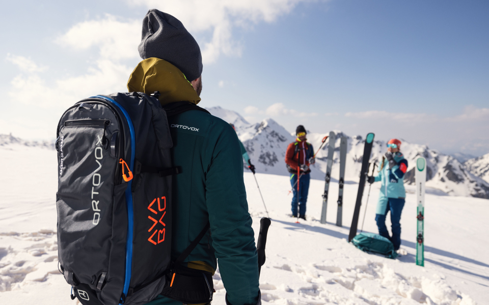 Skifahrer mit Ortovox-Rucksack auf schneebedecktem Berggipfel, mit Skifahrern und Skiern im Hintergrund.