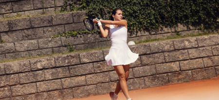 Frau im weißen Kleid spielt Tennis auf einem Sandplatz und schwingt den Schläger, um den Ball zu schlagen.
