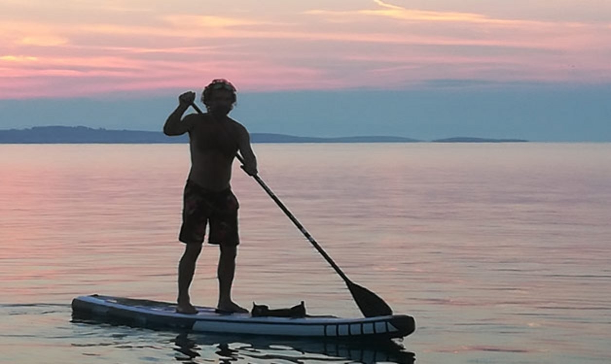Mann steht beim Sonnenuntergang auf einem Paddleboard auf ruhigem Meerwasser; Stand-Up-Paddleboarding-Fitness.