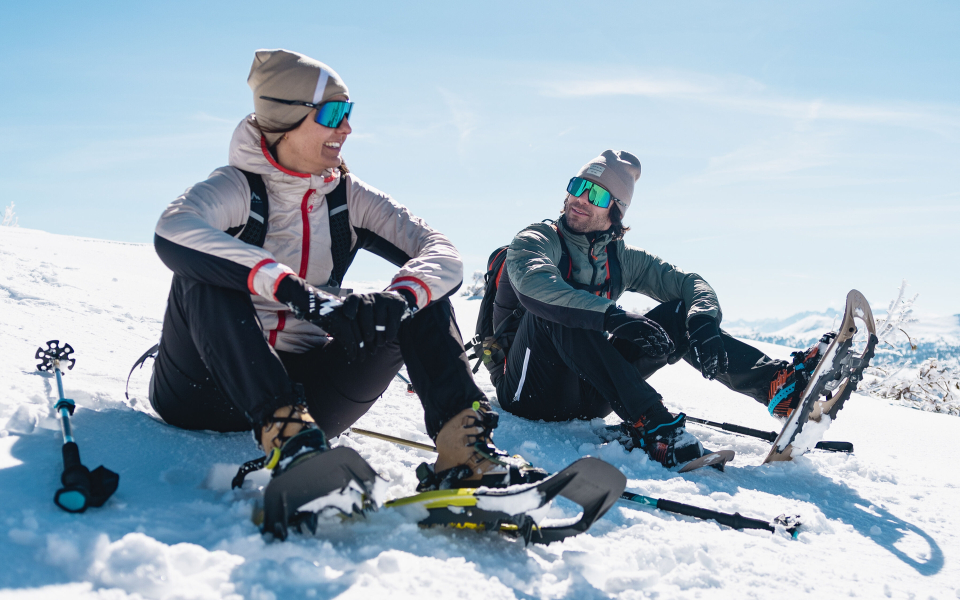 Zwei Freunde machen eine Schneeschuhwanderung und ruhen sich an einem sonnigen Wintertag im Schnee aus, mit Schneeschuhen und Stöcken.