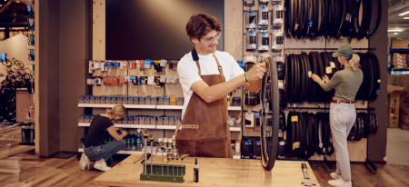Bike shop: Mechanic repairs bicycle wheel as customers browse tires and accessories.
