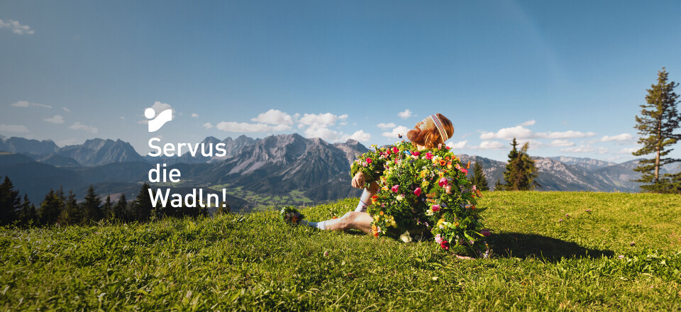 Frau, die in einem Park läuft und ein geblümtes Kleid trägt, mit dem Text "Servus die Wadln!" als Overlay.