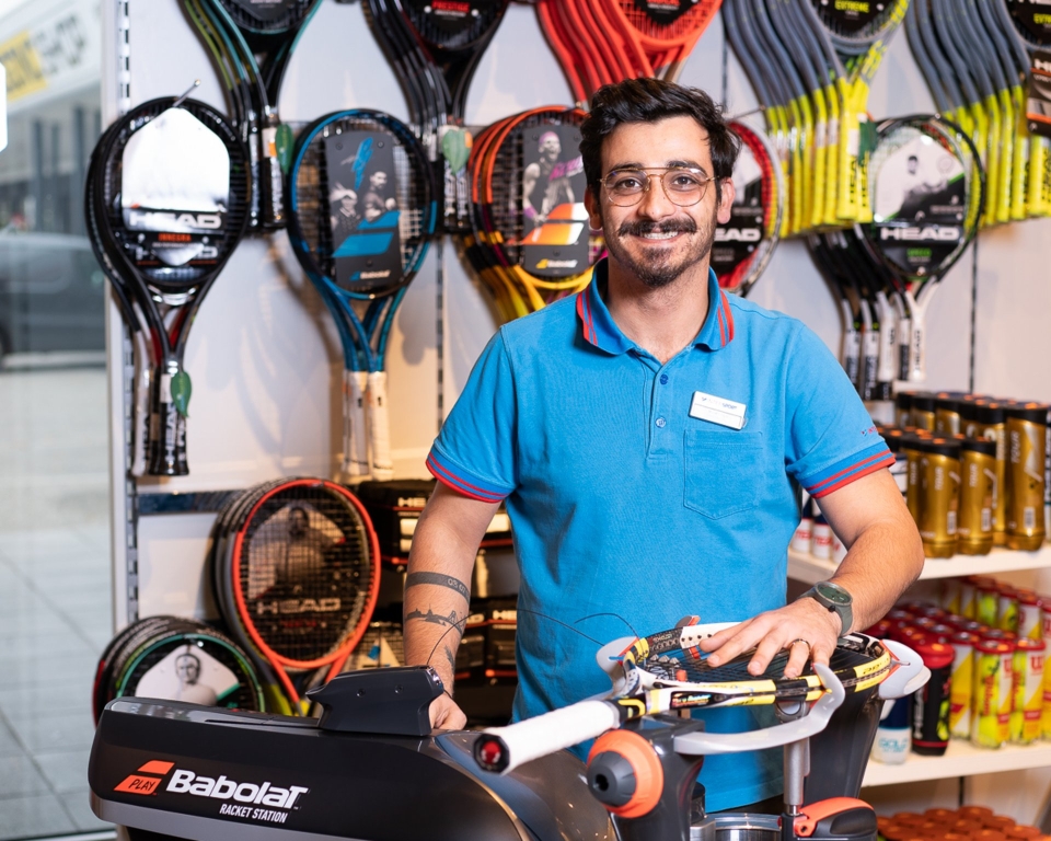 Tennisgeschäft-Mitarbeiter mit Besaitungsmaschine, umgeben von Tennisschlägern und Ausrüstung.