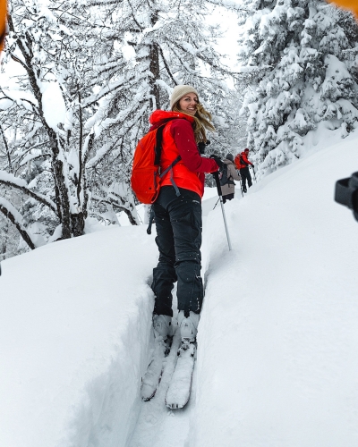 Frau beim Tiefschneefahren im Backcountry, mit roter Jacke und Rucksack, winterliche Berglandschaft.