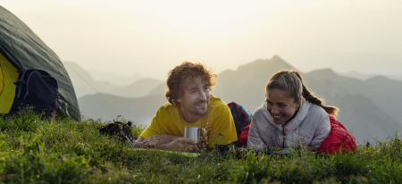 Paar campt in den Bergen und entspannt sich draußen vor dem Zelt in Schlafsäcken mit malerischer Aussicht.