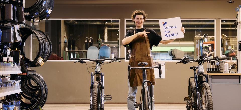 Ein Fahrradladenmitarbeiter steht mit einem "Service de Waffin"-Schild zwischen Mountainbikes in einem Einzelhandelsgeschäft.