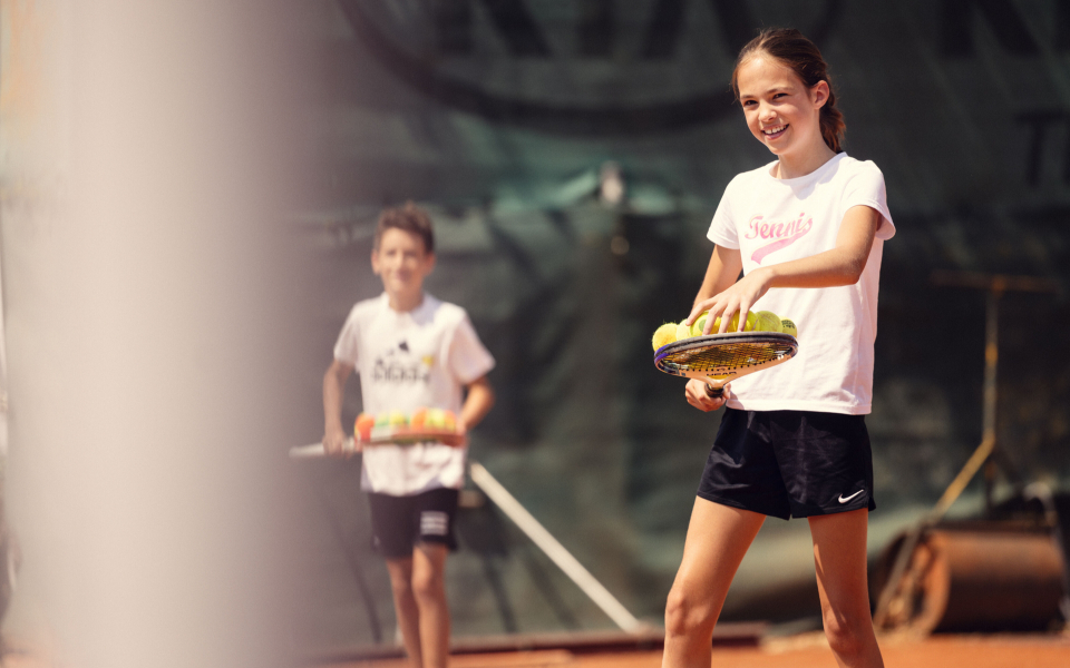 Junges Mädchen balanciert Tennisbälle auf einem Schläger während einer Tennisstunde für Kinder mit einem Trainer.