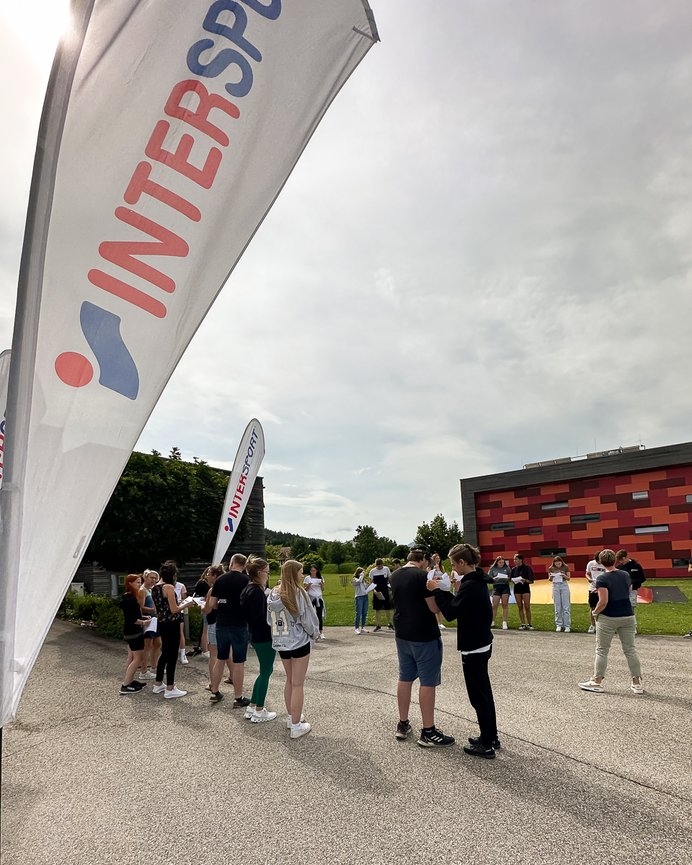 Eine Gruppe von Menschen im Freien in der Nähe einer Intersport-Flagge, möglicherweise bei einer Sportveranstaltung oder einer Geschäftseröffnung.
