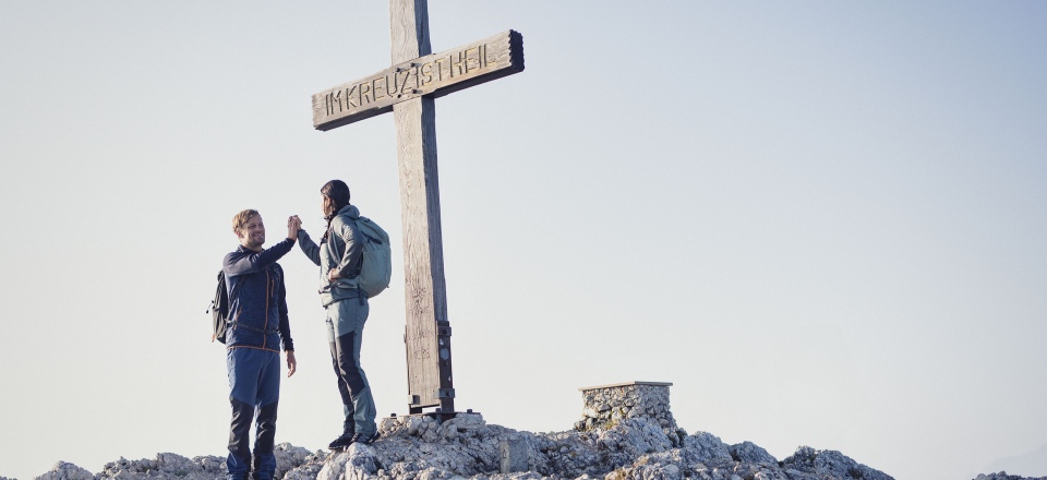 Zwei Wanderer geben sich an einem Gipfelkreuz ein High-Five, um eine erfolgreiche Bergwanderung zu feiern.