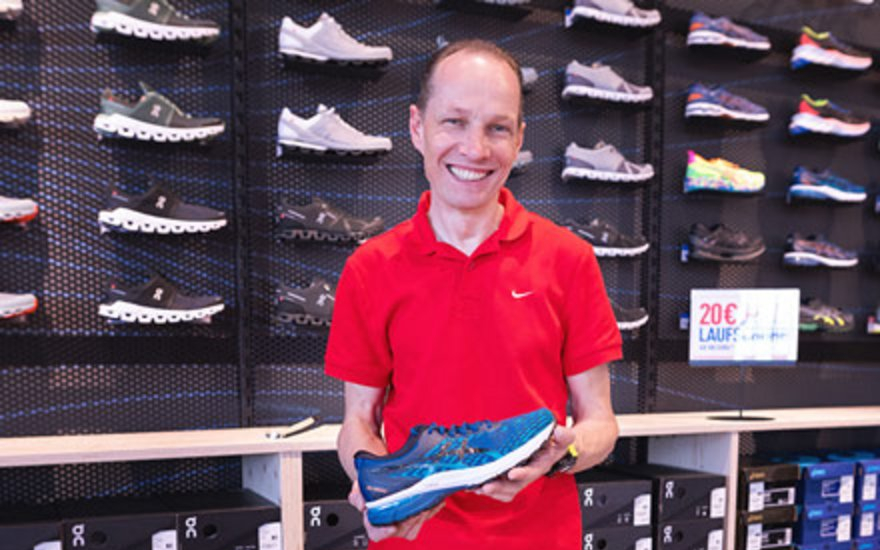 Mann im roten Hemd hält einen blauen Laufschuh in einem Geschäft mit einer Auslage für Sportschuhe.