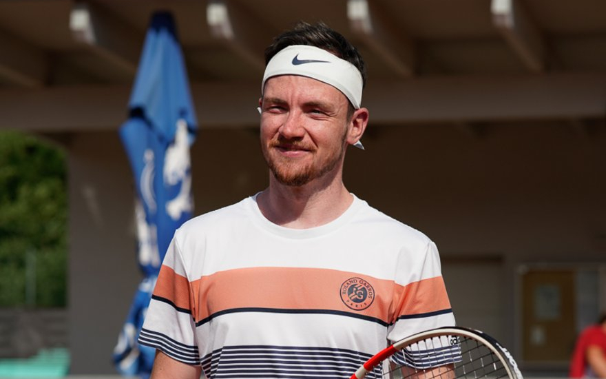 Lächelnder Tennisspieler mit Schläger und Ball auf einem Sandplatz, der ein Nike-Stirnband trägt.