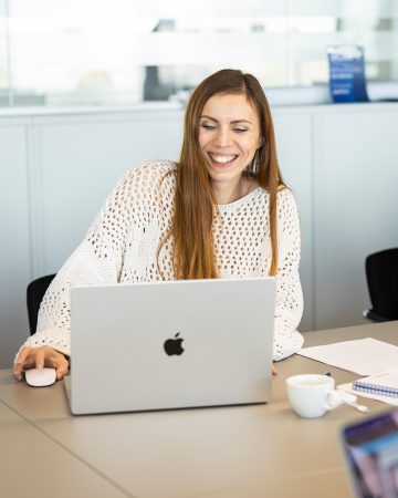 Lächelnde Frau arbeitet an einem Laptop in einem hellen Büro mit Kaffee und Dokumenten auf dem Schreibtisch.