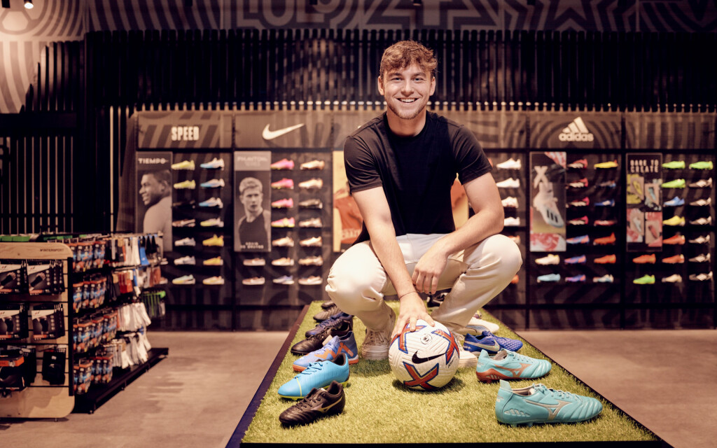 Lächelnder Mann in einem Sportgeschäft mit Fußball und Fußballschuhen; Nike, Adidas Schuh-Auslage.