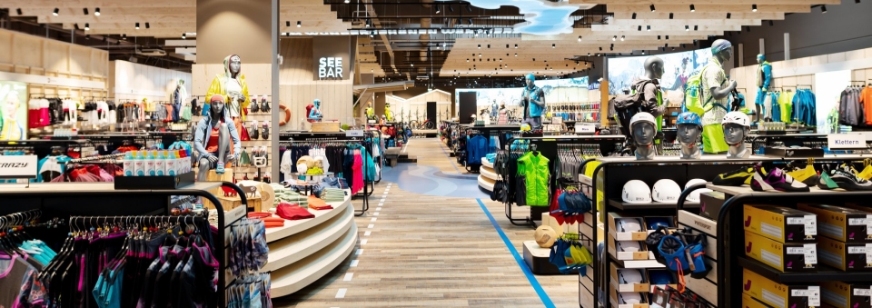 Innenansicht eines Outdoor-Ausrüstungsgeschäfts mit Auslagen für Kleidung, Schuhe und Ausrüstung auf Regalen und Gestellen.