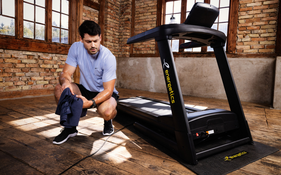 Mann wischt sich nach dem Training auf einem Energetics-Laufband in einem Home-Gym mit Ziegelwand und Holzböden den Schweiß ab.