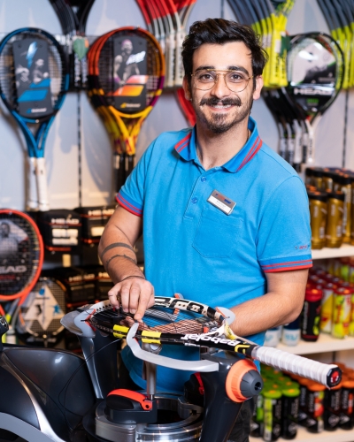 Lächelnder Mann mit Tennisschläger in einem Sportgeschäft, umgeben von Tenniszubehör und Schlägern.