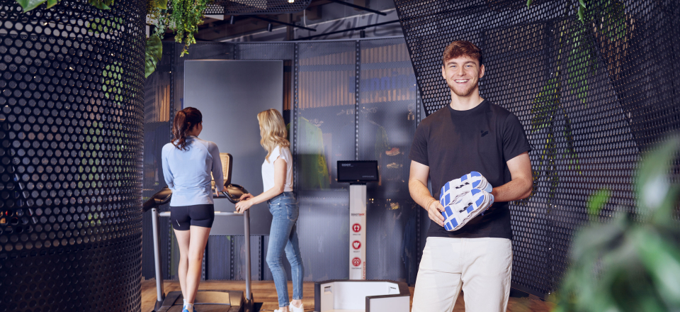 Mann hält ein Paket in einem modernen Fitnessstudio mit Frauen auf einem Laufband und einem Gerät zur Körperanalyse.