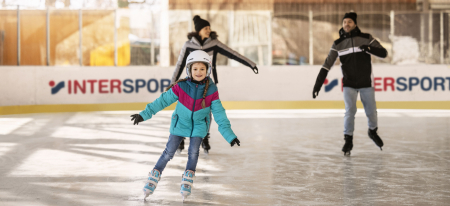 Familieneislaufen in einer Indoor-Eisbahn; Mädchen mit Helm und bunter Jacke geht voran.
