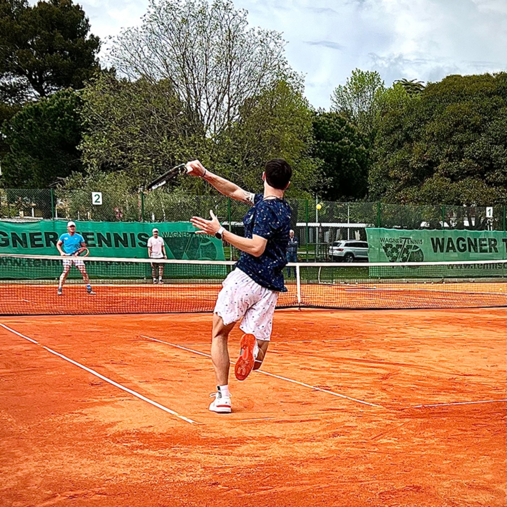 Tennisspieler serviert auf Sandplatz während eines Outdoor-Matches mit Gegner und Bäumen.