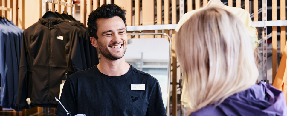 Ein freundlicher Verkäufer hilft einem Kunden bei der Auswahl von Kleidung in einem Outdoor-Bekleidungsgeschäft.