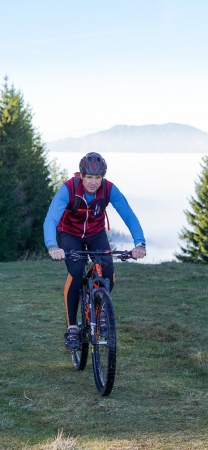 Mann fährt mit dem Mountainbike auf einem grasbewachsenen Hügel mit Wald und Nebel im Hintergrund. Outdoor-Fahrradabenteuer.