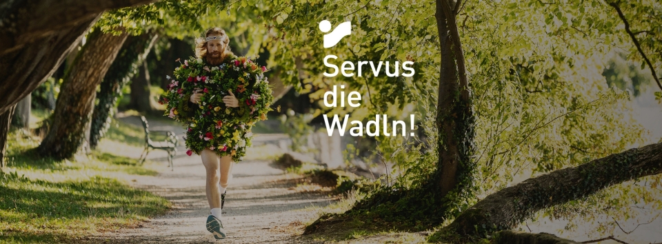 Frau, die in einem Park läuft und ein geblümtes Kleid trägt, mit dem Text "Servus die Wadln!" als Overlay.