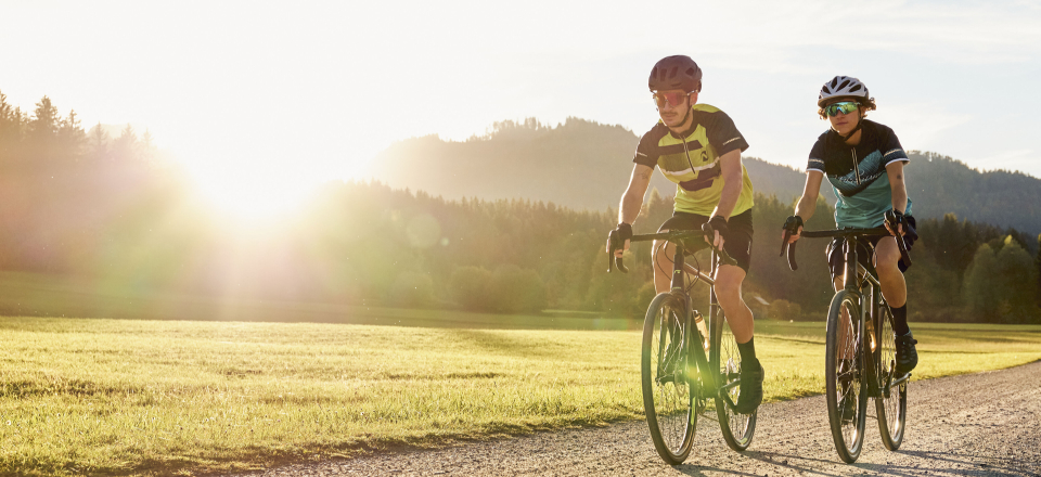 Zwei Radfahrer fahren mit Gravelbikes auf einer unbefestigten Straße durch eine sonnige Wiesenlandschaft.