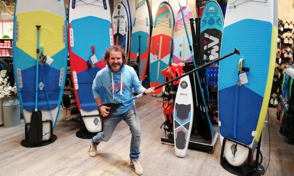 Mann mit Paddel in Surfshop, umgeben von farbenfrohen Stand-Up-Paddle-Boards.